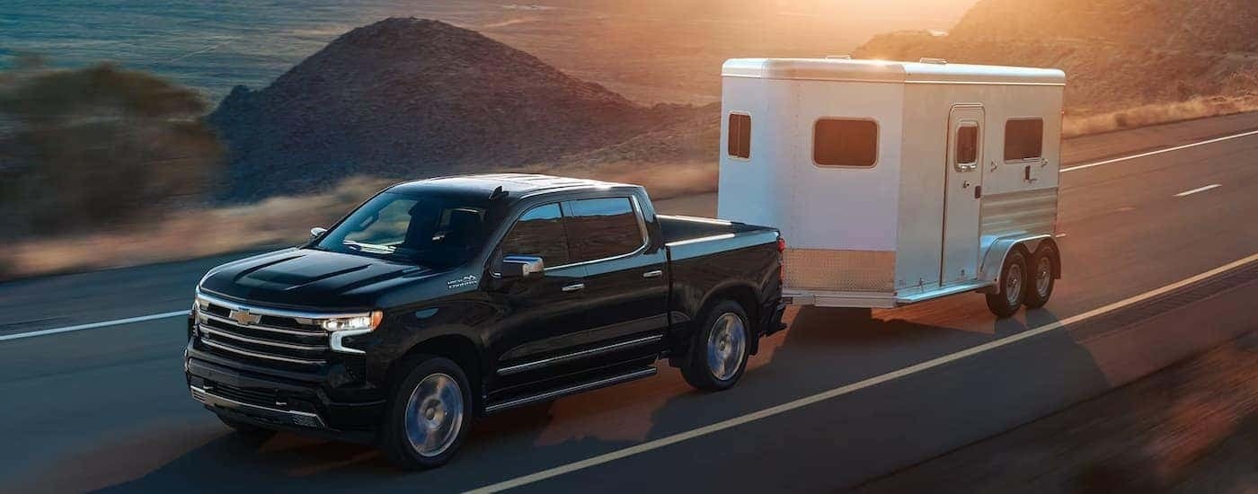 A black 2024 Chevy Silverado 1500 High Country is towing a white trailer on an open road.