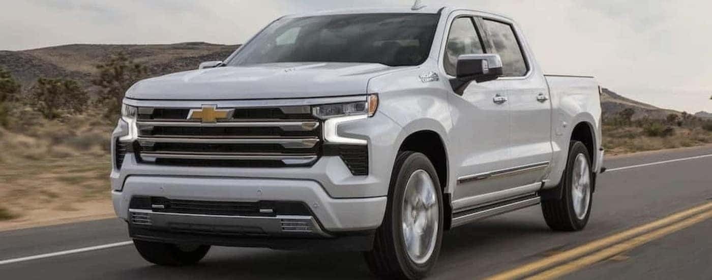 A white 2024 Chevy Silverado 1500 High Country is shown driving on a highway to a Chevy Silverado 1500 dealer