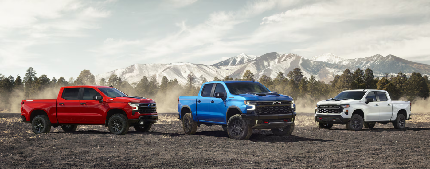 A red, a blue, and a white 2024 Chevy Silverado for sale near Pelham are parked on dirt.